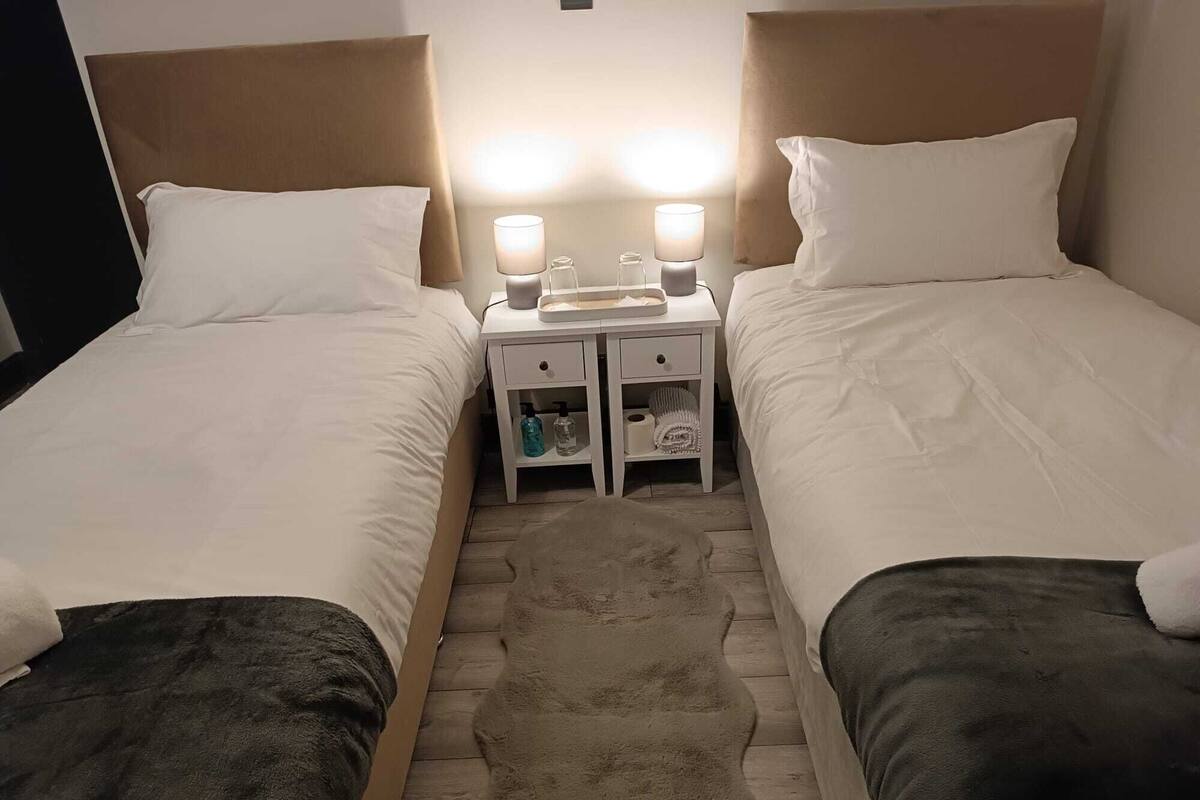A double room with two beds, each with white bedding and a brown headboard. There are two bedside tables with lamps, a small drawer, and a shelf with toiletries.