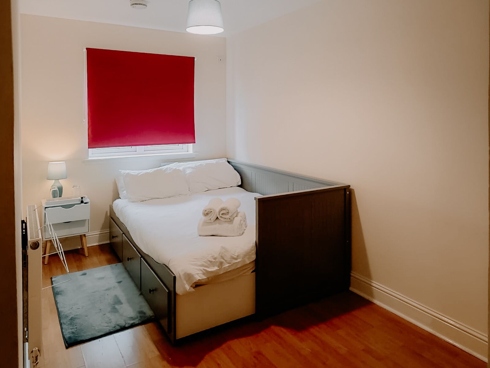 A bedroom with a bed, a bedside table with a lamp, a window with a red blind, and a wooden floor.