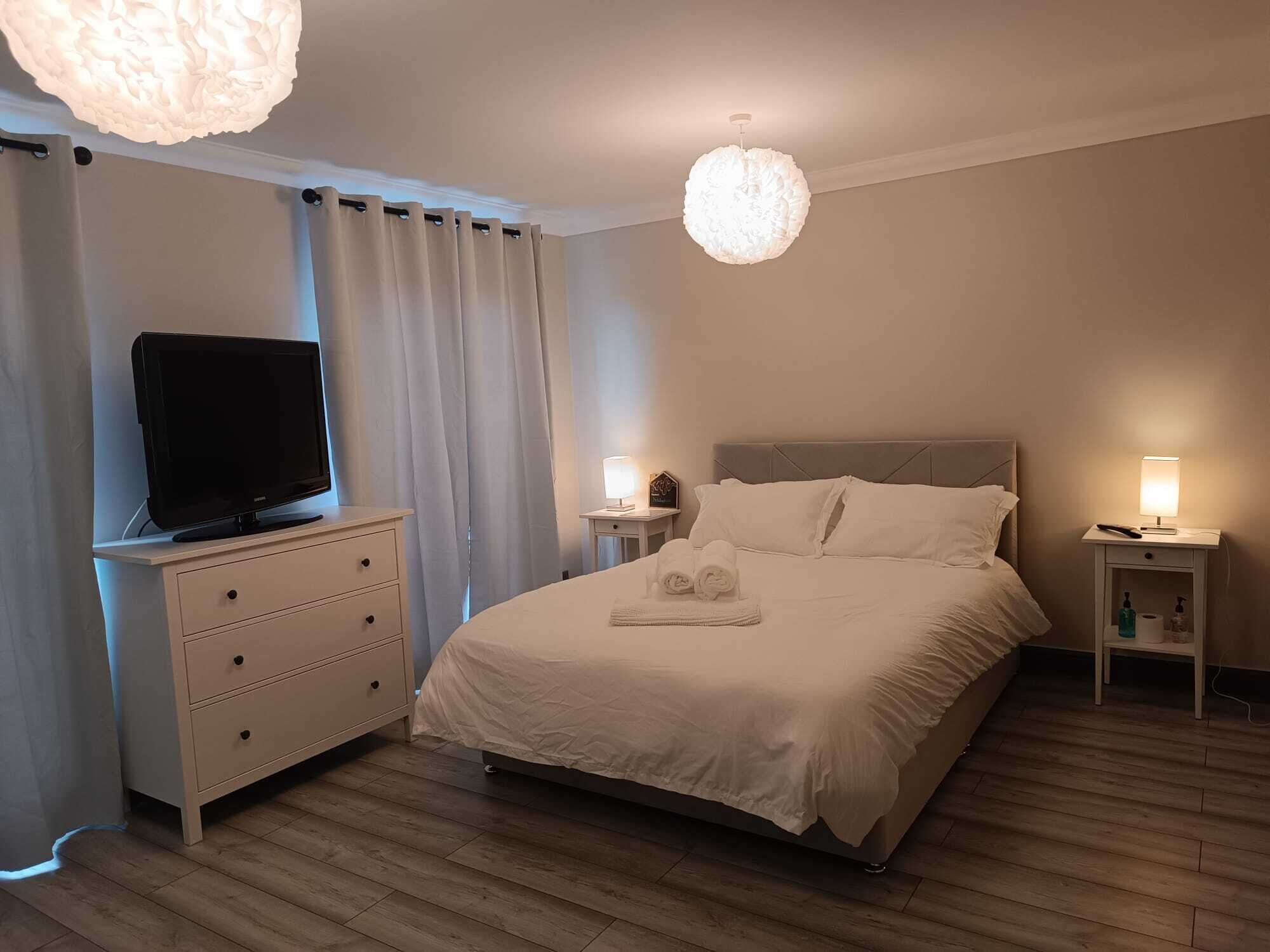 A hotel room with a bed, a TV on a dresser, two bedside lamps, and a large chandelier.