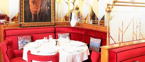 A dining area with red upholstered seating, a white tablecloth, and a large framed painting on the wall.