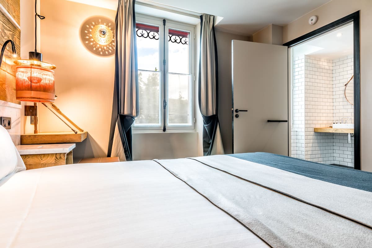 Ein Hotelzimmer mit Bett, Nachttischlampe, einem Fenster mit Vorhängen und einem durchgehenden Türspalt sichtbaren Badezimmer.