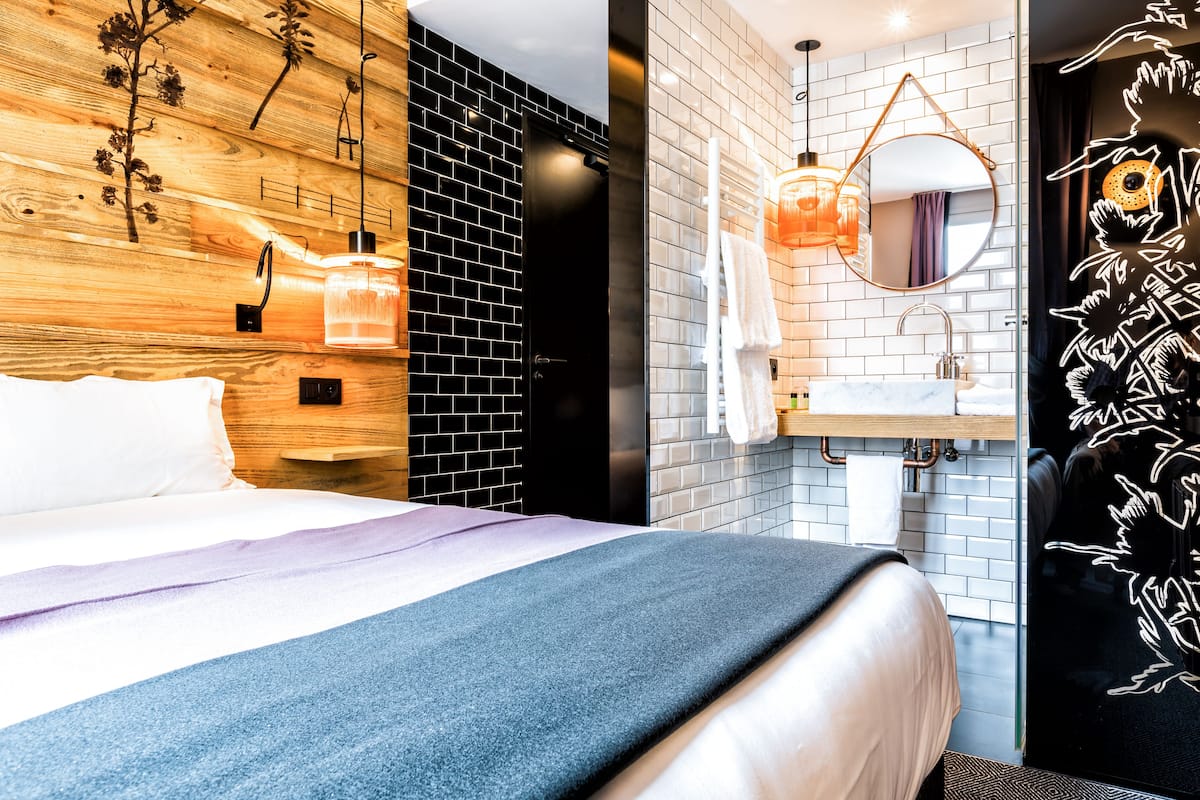 Ein modernes Hotelzimmer mit einer Wand in Holzoptik, einem großen Bett mit blauer Decke, einem Badezimmer mit weißem Waschbecken und Spiegel sowie einer schwarzen Tür mit floralem Dekor.