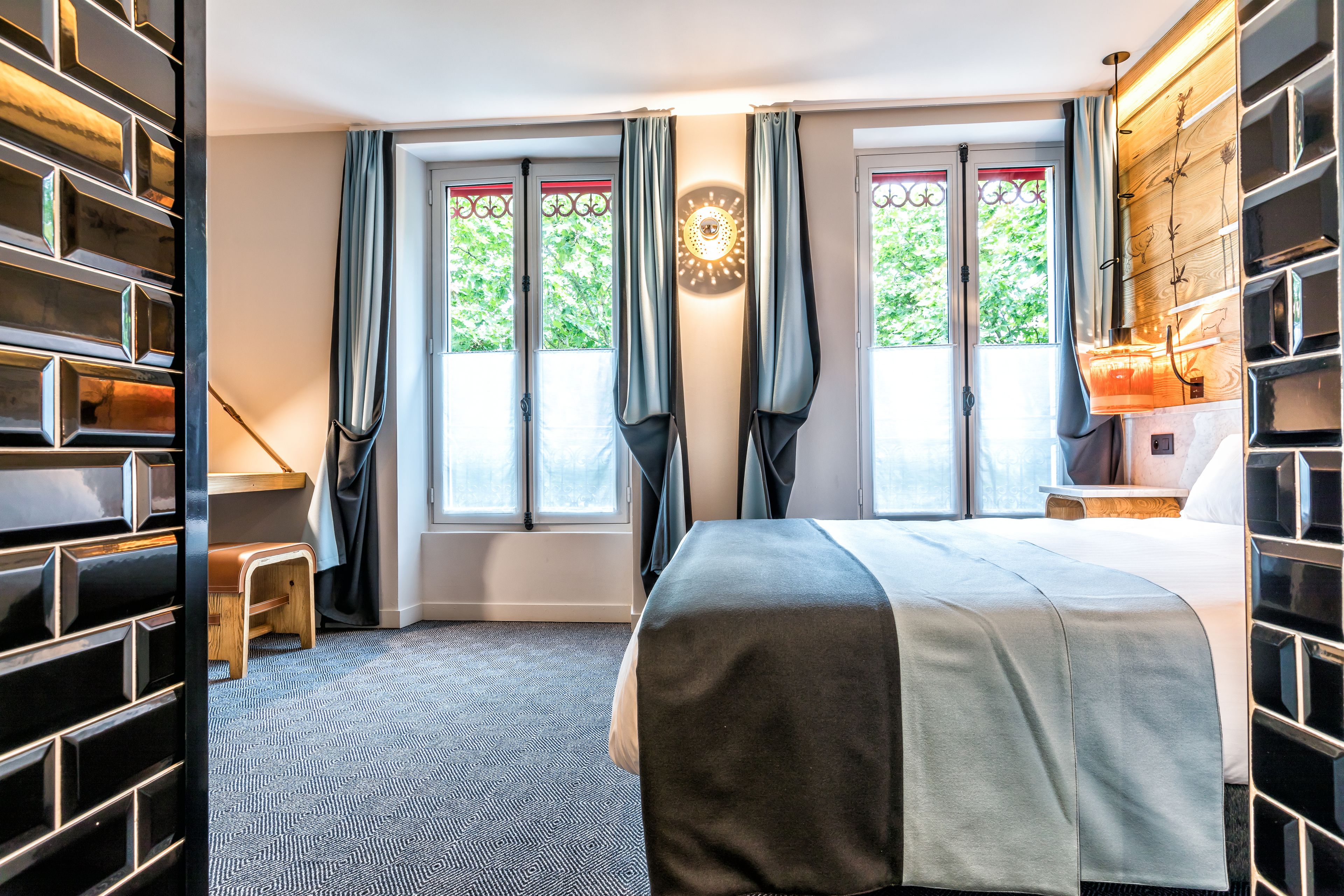 A hotel room with a bed, a small seating area, a window with curtains, and a wall-mounted shelf.