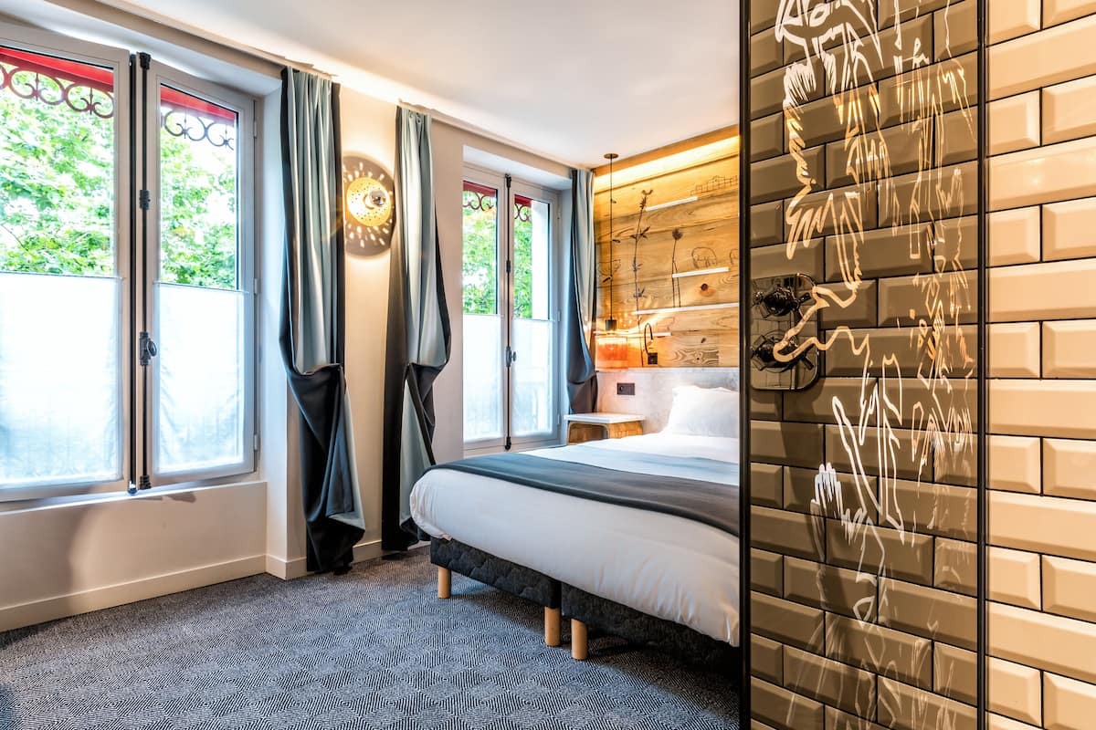 Ein Hotelzimmer mit Bett, einer Wand aus Holzpaneelen und Blick auf Bäume durch die Fenster.