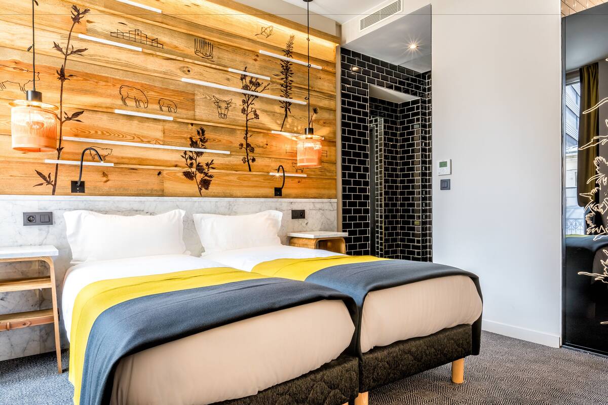 Ein modernes Hotelzimmer mit einer Wand in Holzoptik, einem Bett mit blauer und gelber Bettdecke sowie einem Duschbereich mit schwarz-weißen Fliesen.