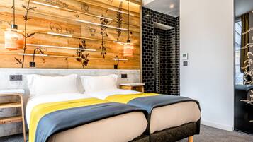 A modern hotel room with a wooden accent wall, a bed with a yellow and blue bedspread, and a black and white tiled shower area.