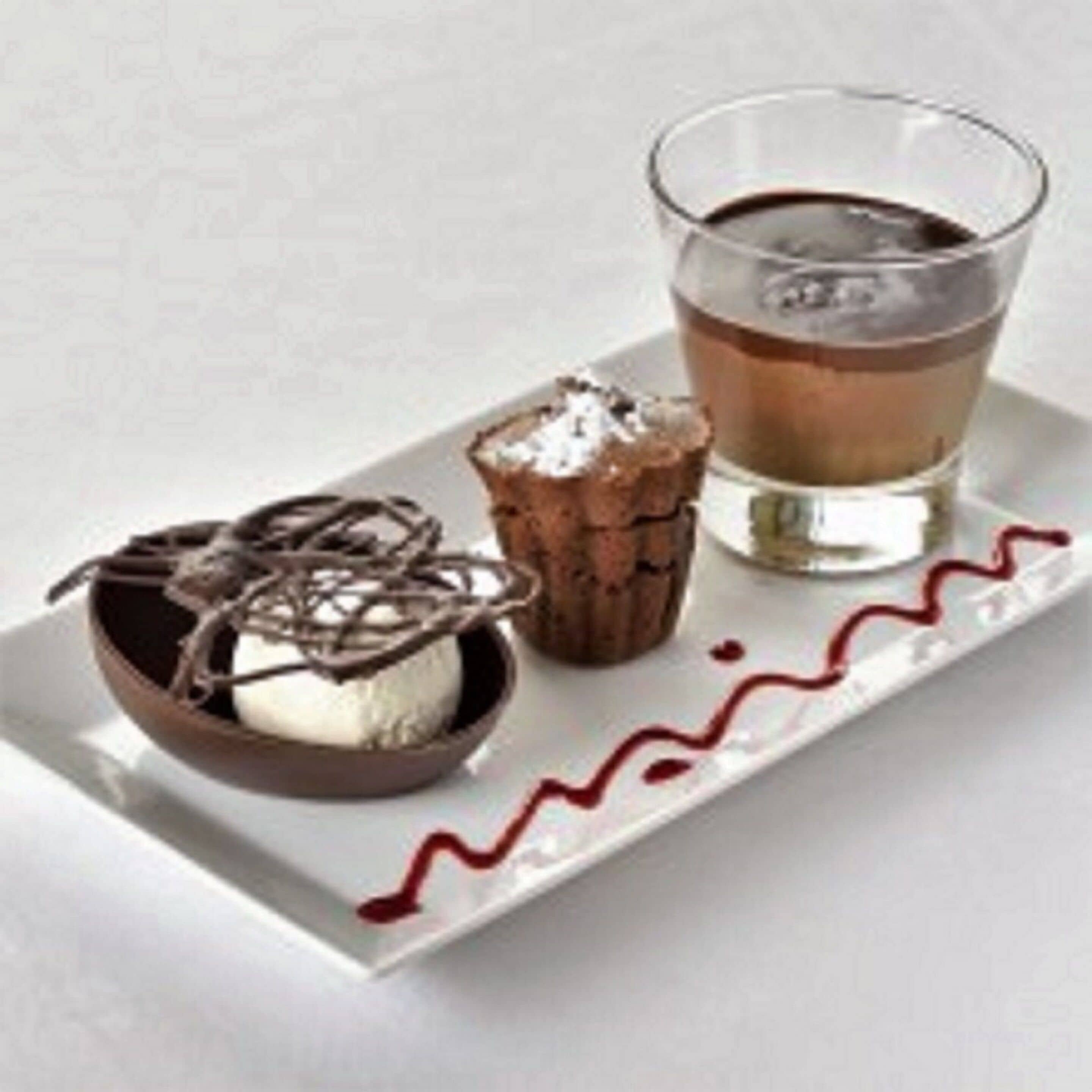 A dessert tray with a chocolate mousse, a small cupcake, and a glass of dark liquid, possibly coffee or tea, on a white plate with a red wavy line design.