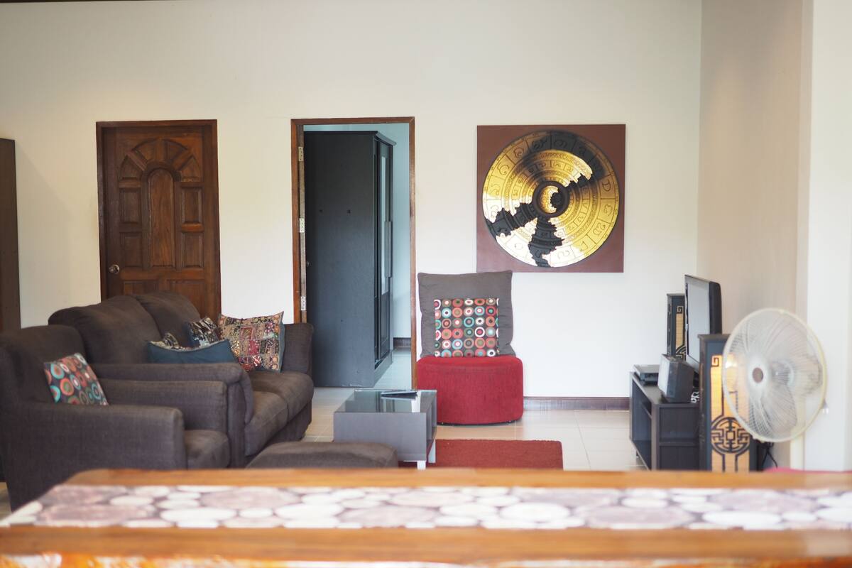 Un salon comprenant un canapé, un pouf rouge, une table basse, un meuble pour la télévision et un ventilateur fixé au mur.