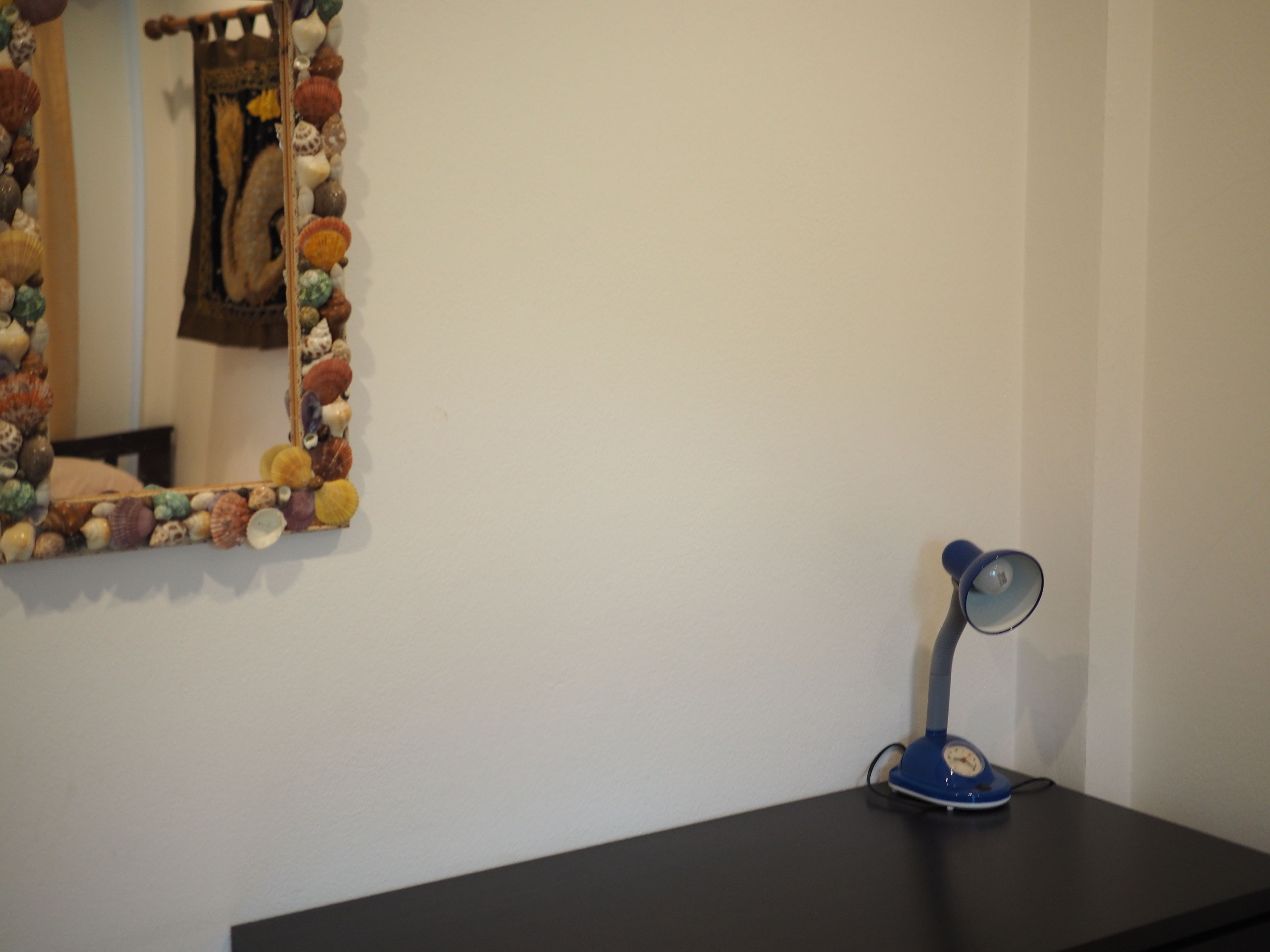 Un miroir avec un cadre en forme de coquillage, une lampe de bureau bleue et un bureau noir, le tout placé devant un mur blanc.