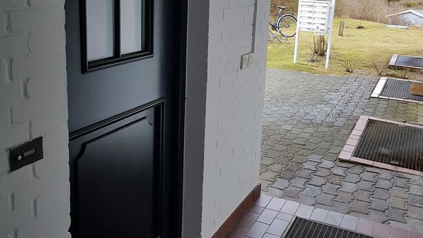 A black door with a window, a white brick wall, and a paved entrance area with a metal grate. - Haus Stedum - App. Lenz (Sylt)