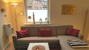 A living room with a grey sofa, red cushions, a white coffee table, and a window with a view of buildings. - Haus Stedum - App. Lenz (Sylt)