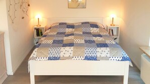 A bedroom with a white bed, patterned bedding, two bedside tables with lamps, a wall-mounted picture, and a decorative wall piece. - Haus Stedum - App. Lenz (Sylt)