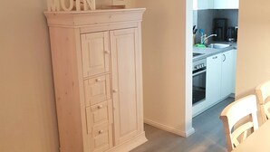 A room with a white cabinet, a table with chairs, and a kitchen area visible through an open doorway. - Haus Stedum - App. Lenz (Sylt)