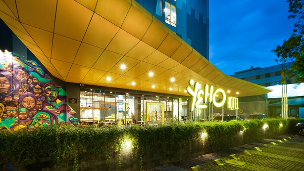 Een hotelentree met een groot verlicht bord, een modern architectonisch ontwerp en een goed verlicht buitenterrein met zitplaatsen.