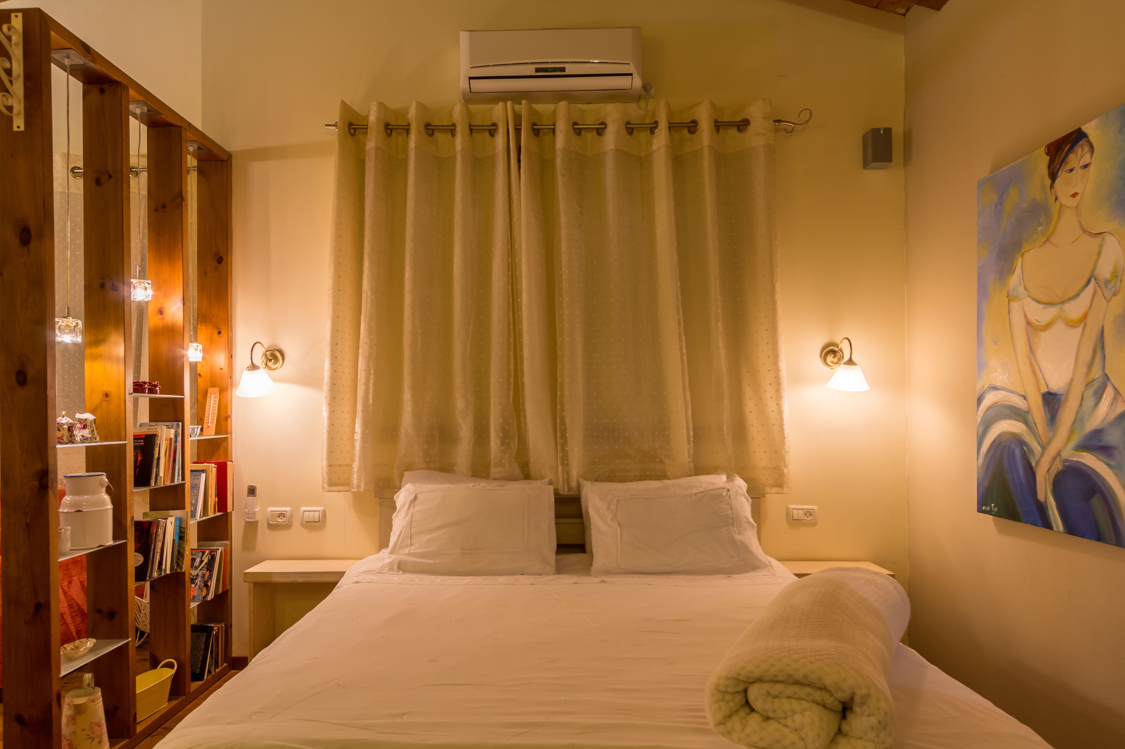 A hotel room with a bed, a bookshelf, an air conditioning unit, and a painting on the wall.
