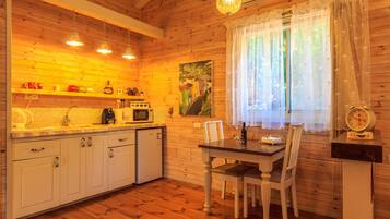 A cozy wooden cabin interior with a kitchen featuring white cabinets, a dining table with chairs, and a window with sheer curtains.
