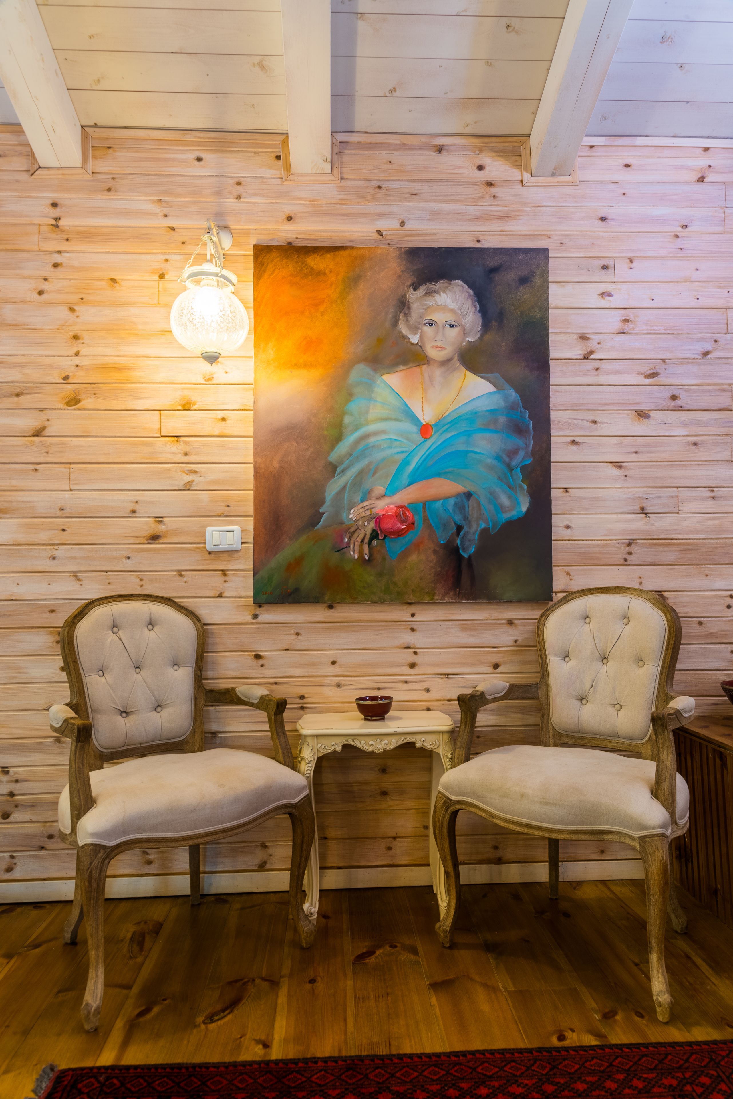 A room with wooden panel walls, two upholstered chairs, a small table, and a painting of a woman holding a red rose.