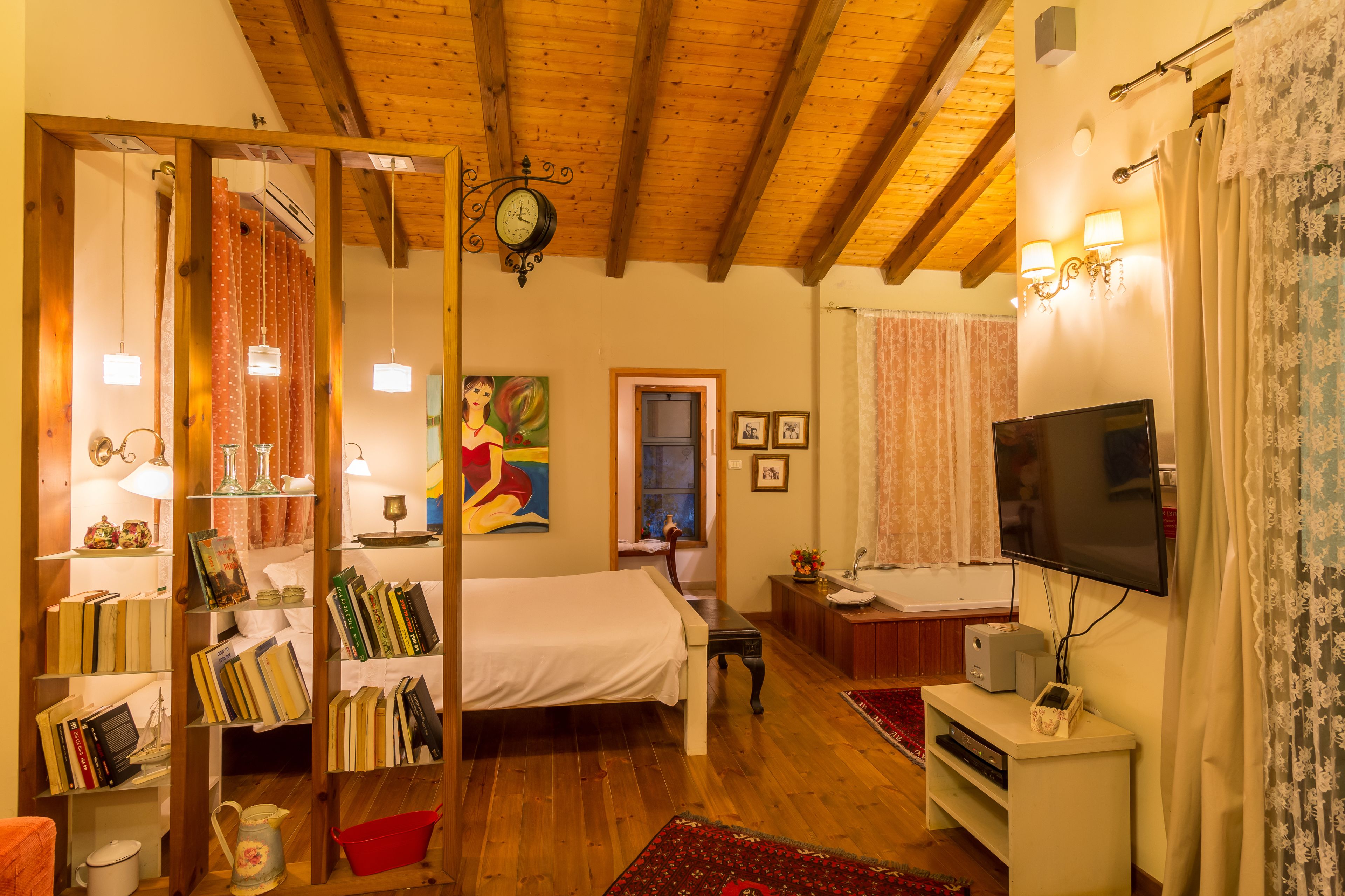 A cozy bedroom with a wooden ceiling, a bed, a bookshelf, a TV, and a bathroom with a shower curtain.
