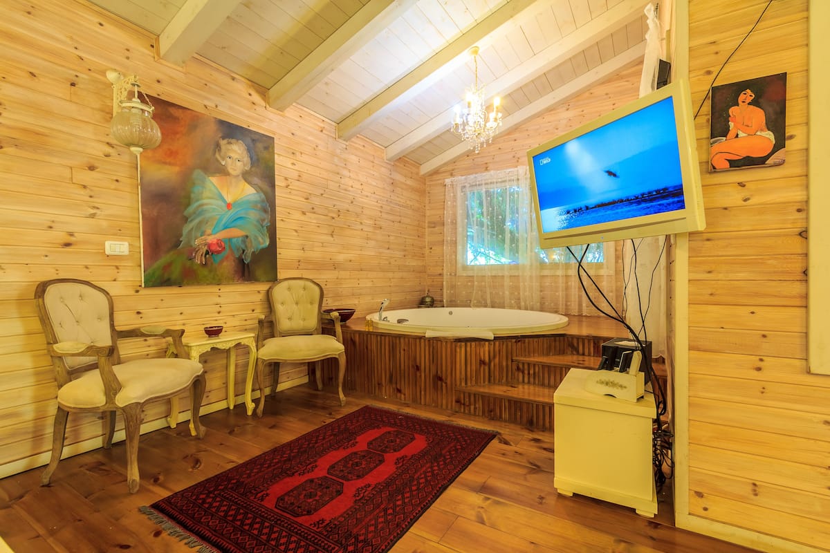 A wooden cabin interior with a large painting, a television, a hot tub, and a red patterned rug.