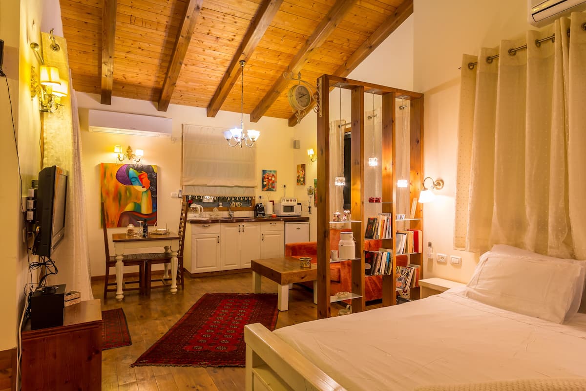 A bedroom with a large bed, a wooden ceiling, a kitchen area, and a dining table.