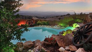 A swimming pool with a wooden fence and a wooden cart wheel, surrounded by rocks and plants. - Betzel Haella (Mevoot HaHermon)