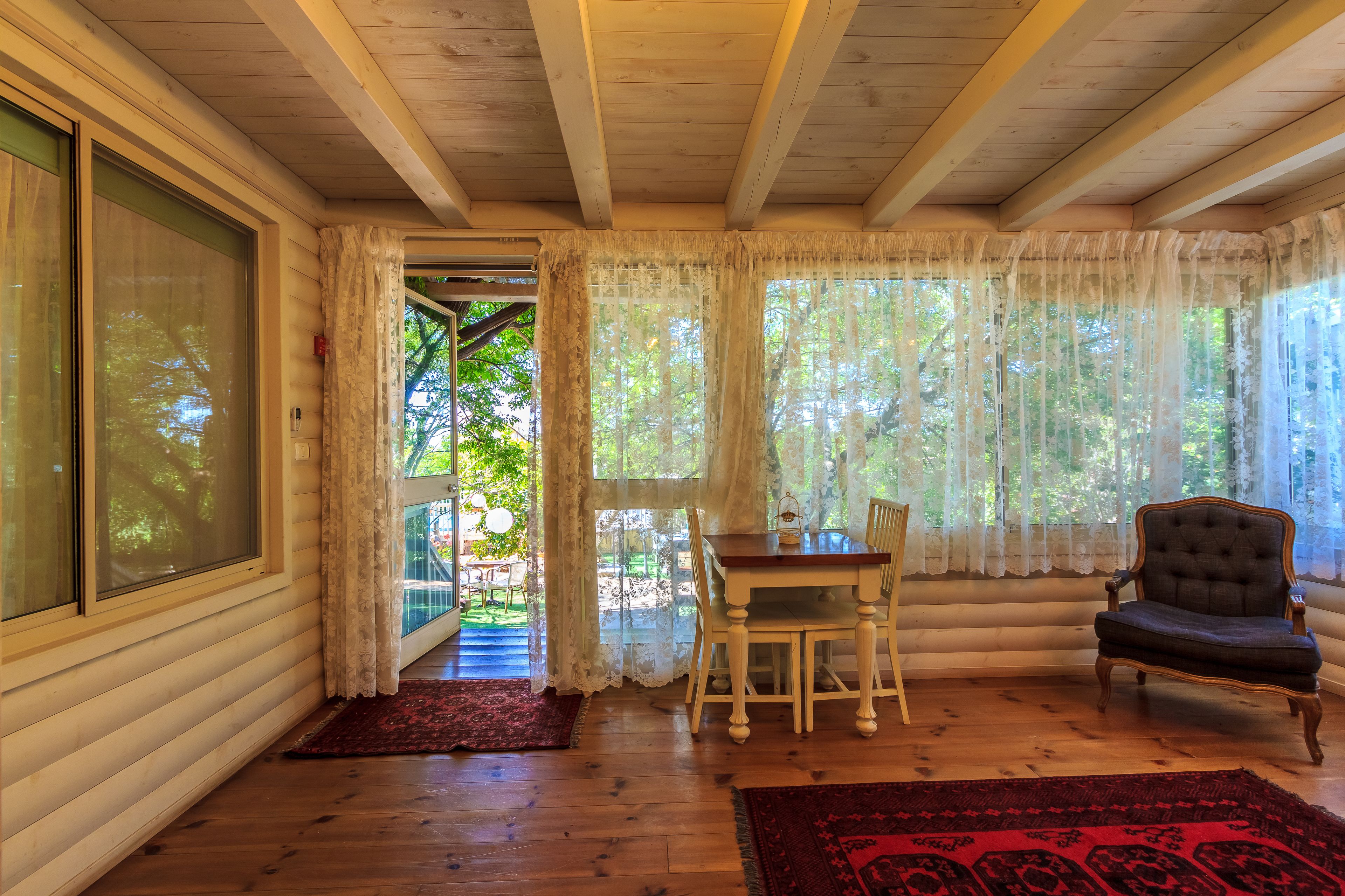 Una habitación acogedora con techo de madera, una mesa de comedor con sillas, un ventanal con cortinas translúcidas y vistas al verdor exterior.