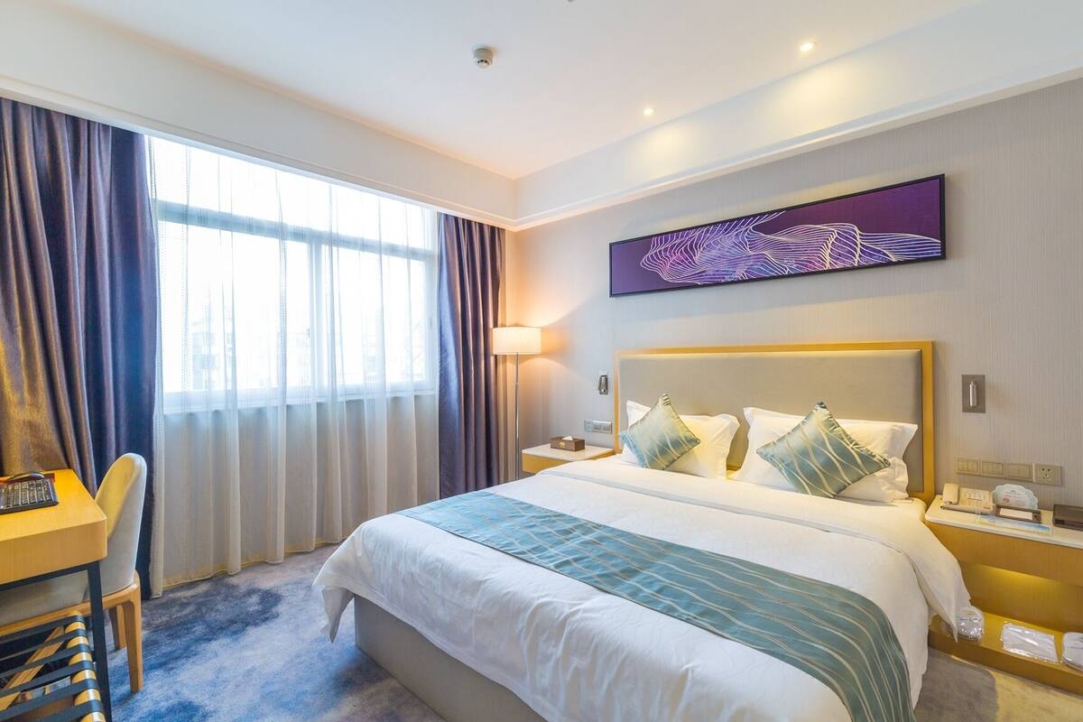 Habitación de hotel con una cama grande, un escritorio y una silla. Se ve una ventana con cortinas, un televisor en la pared y una mesita de noche con una lámpara.