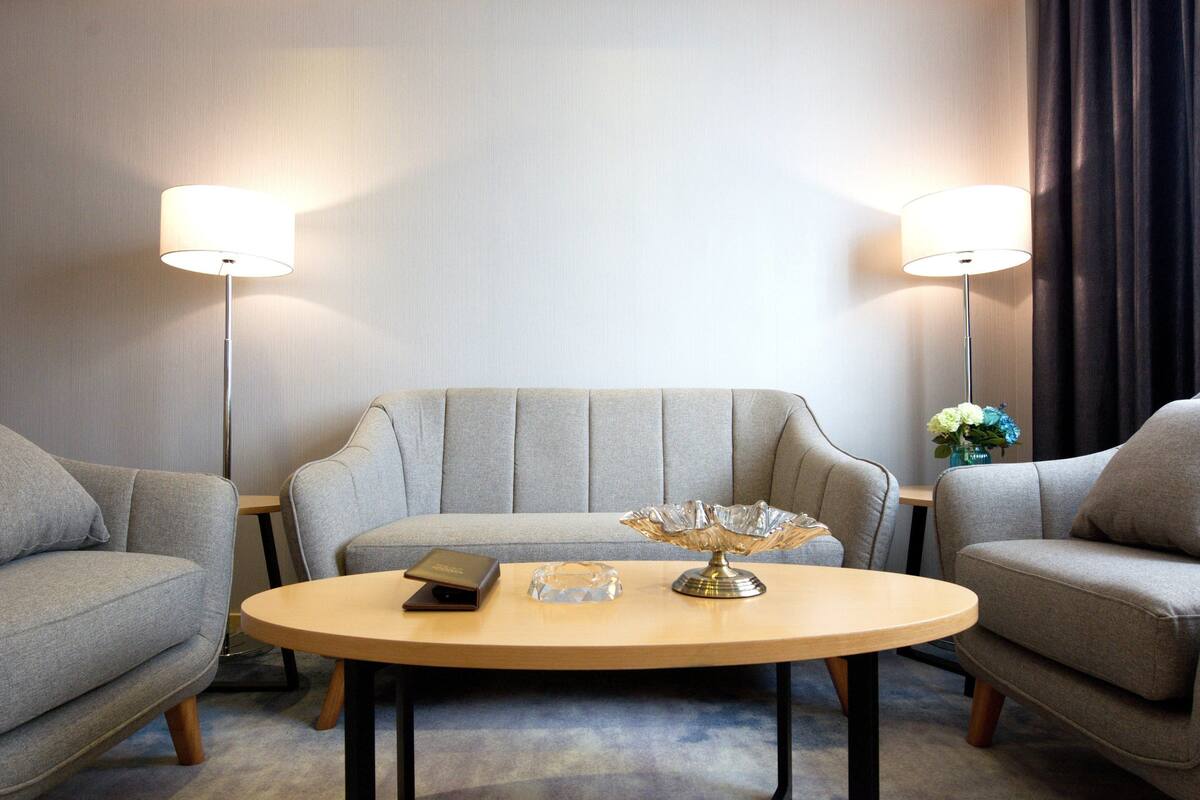 Una habitación de hotel moderna con un sofá gris, una mesa de centro redonda de madera y dos lámparas de pie.