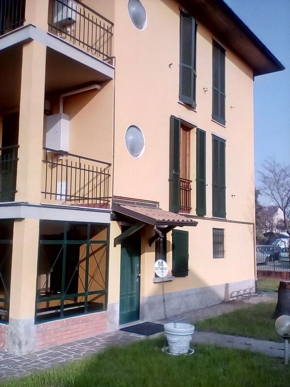 A two-story building with a balcony, a door with a sign, and a small garden area with a fountain. - Locanda I Tarocchi (Vailate)