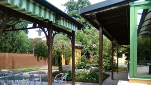 Um pátio externo com mesas e cadeiras, uma área de estar coberta e um jardim com plantas e uma pequena construção.