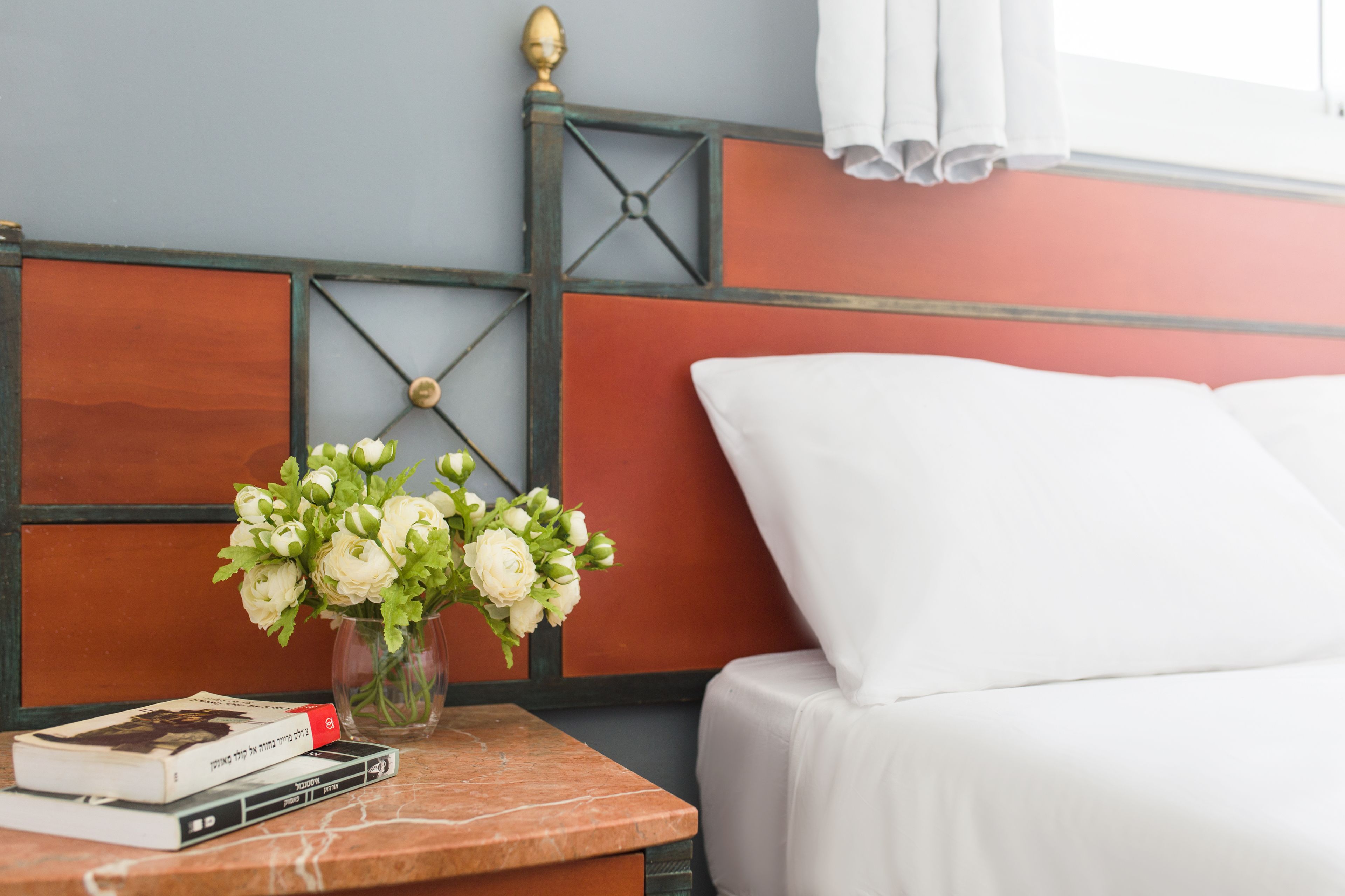 Cama bem arrumada com lençóis brancos, mesa de cabeceira com um livro e um vaso de flores, e parede com design geométrico e janela com cortinas.