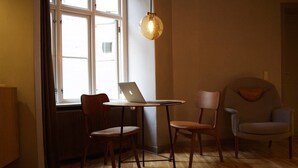 A room with a wooden floor, a round table with a laptop, two wooden chairs, a round armchair, and a hanging light fixture.
