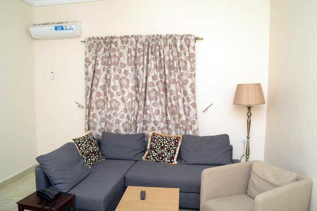 A living room with a grey sofa, two patterned throw pillows, a remote control on a small table, and a lamp.
