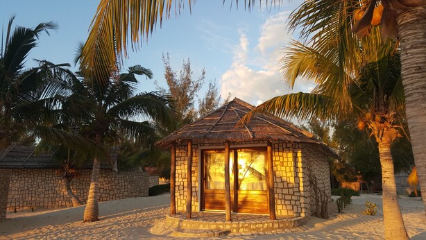 A small stone house with a thatched roof, surrounded by palm trees and a sandy beach. - Laguna Blu - Resort Madagascar (Andavadoaka)
