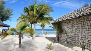 A beach scene with palm trees, a thatched-roof building, and a clear view of the ocean. - Laguna Blu - Resort Madagascar (Andavadoaka)