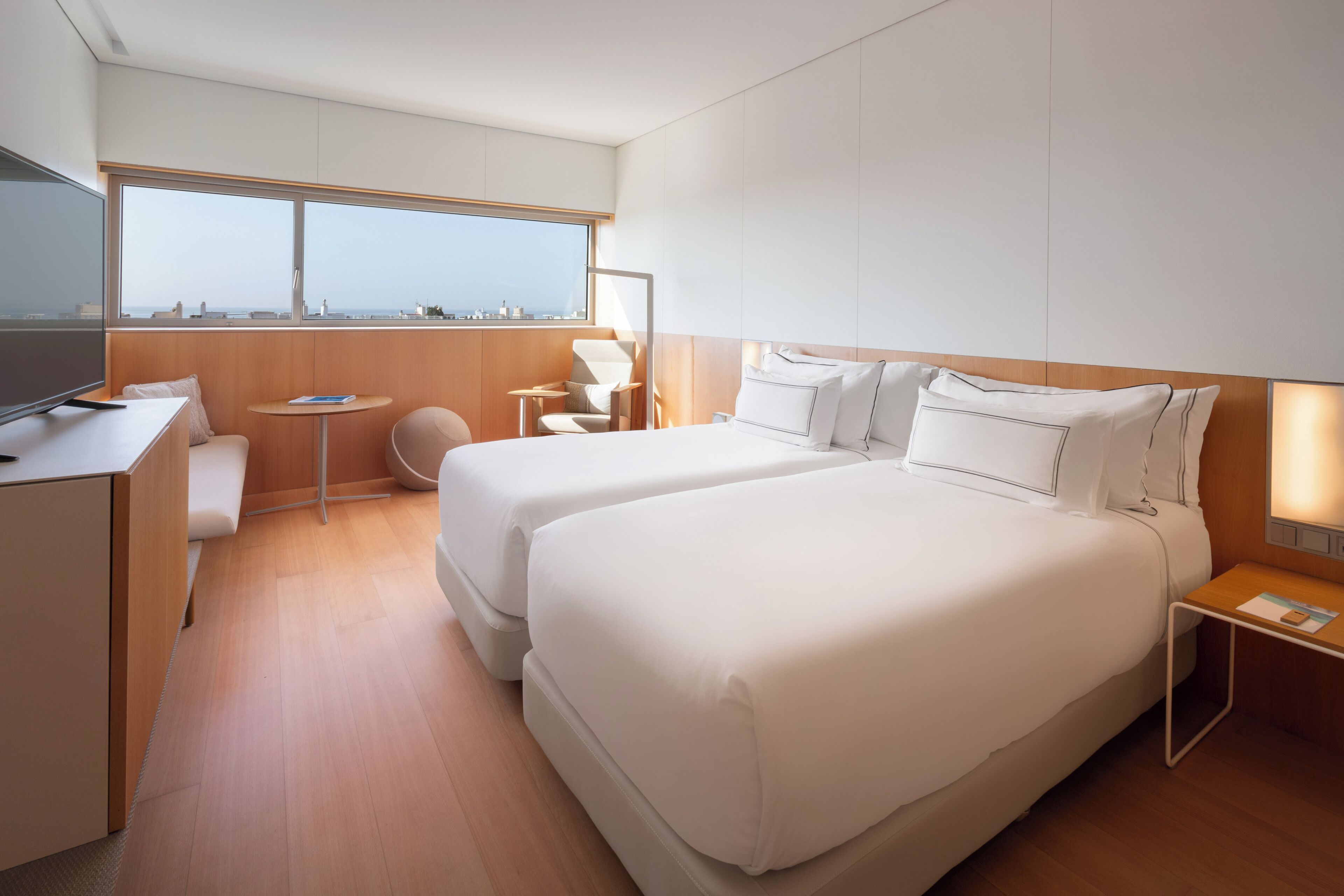 A modern hotel room with two beds, a desk, a chair, a small table, and a large window offering a city view.