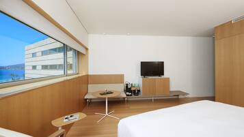 A modern hotel room with a large bed, a TV, a small table with drinks, and a view of the sea.