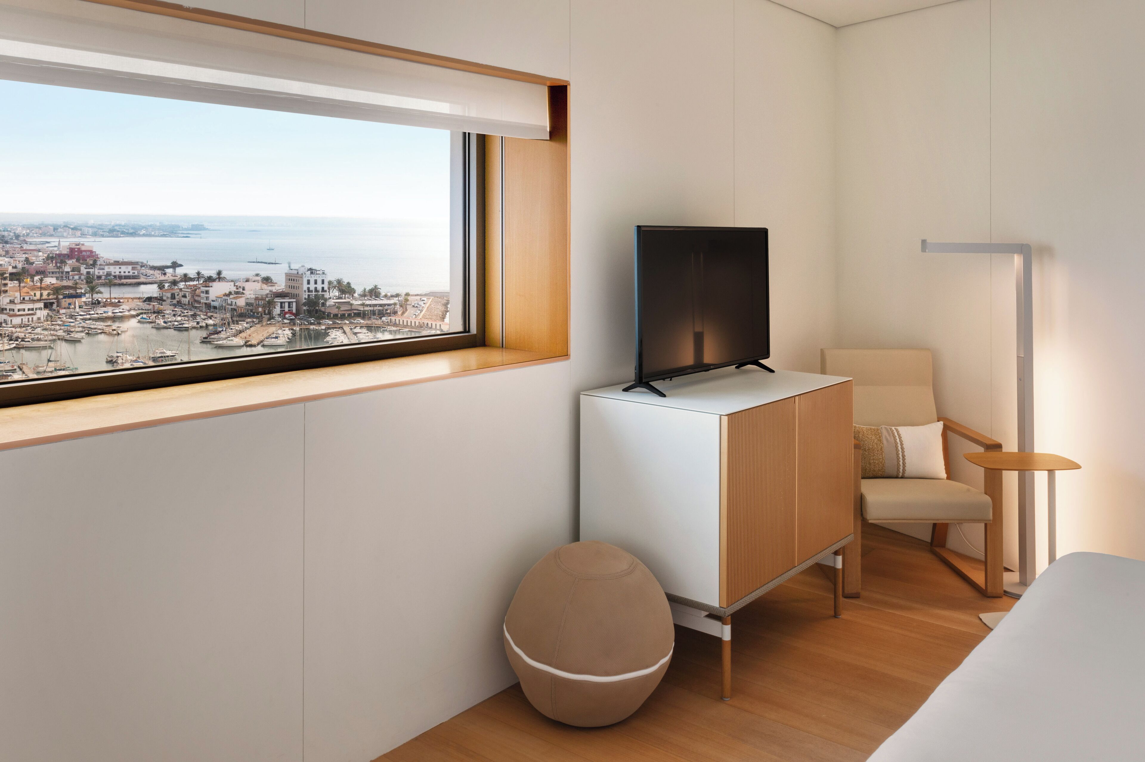 A modern hotel room with a city view, a flat-screen TV, a comfortable chair, a bedside table, and a small ottoman.