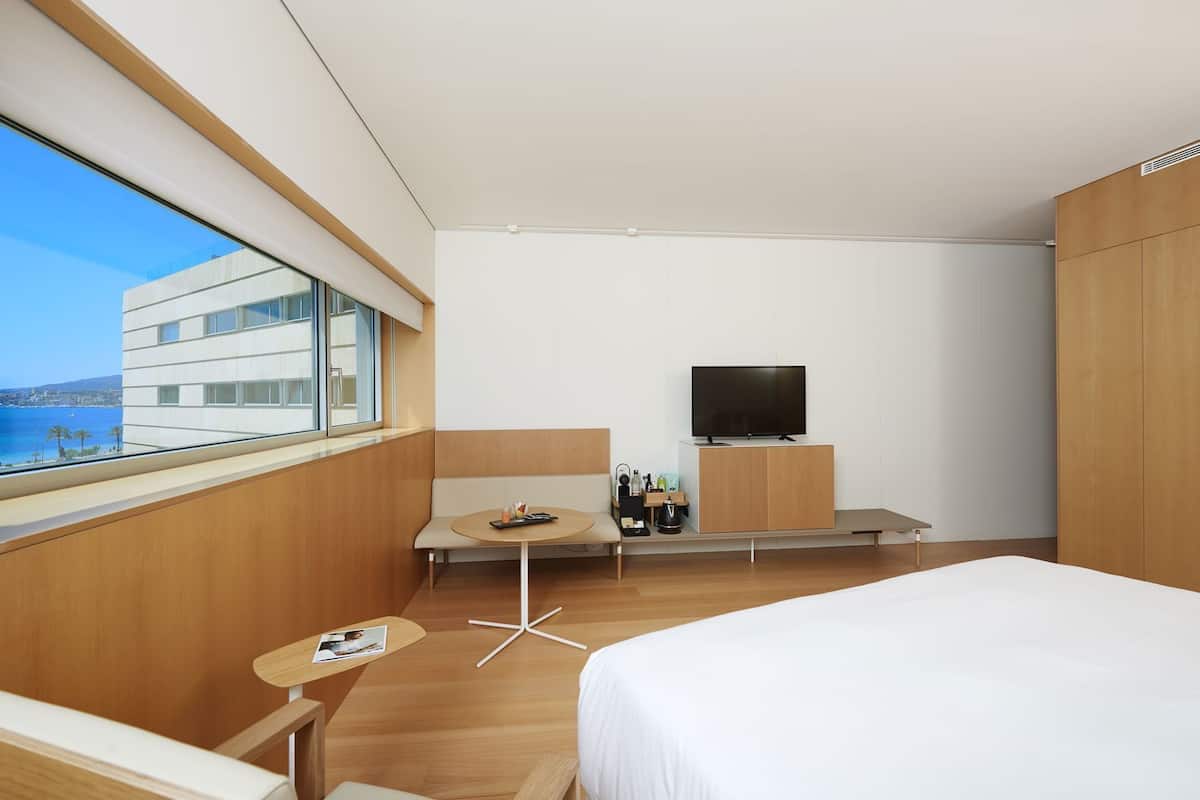 Quarto de hotel moderno com uma cama grande, uma televisão, uma mesa pequena com uma revista e vista para o mar pela janela.