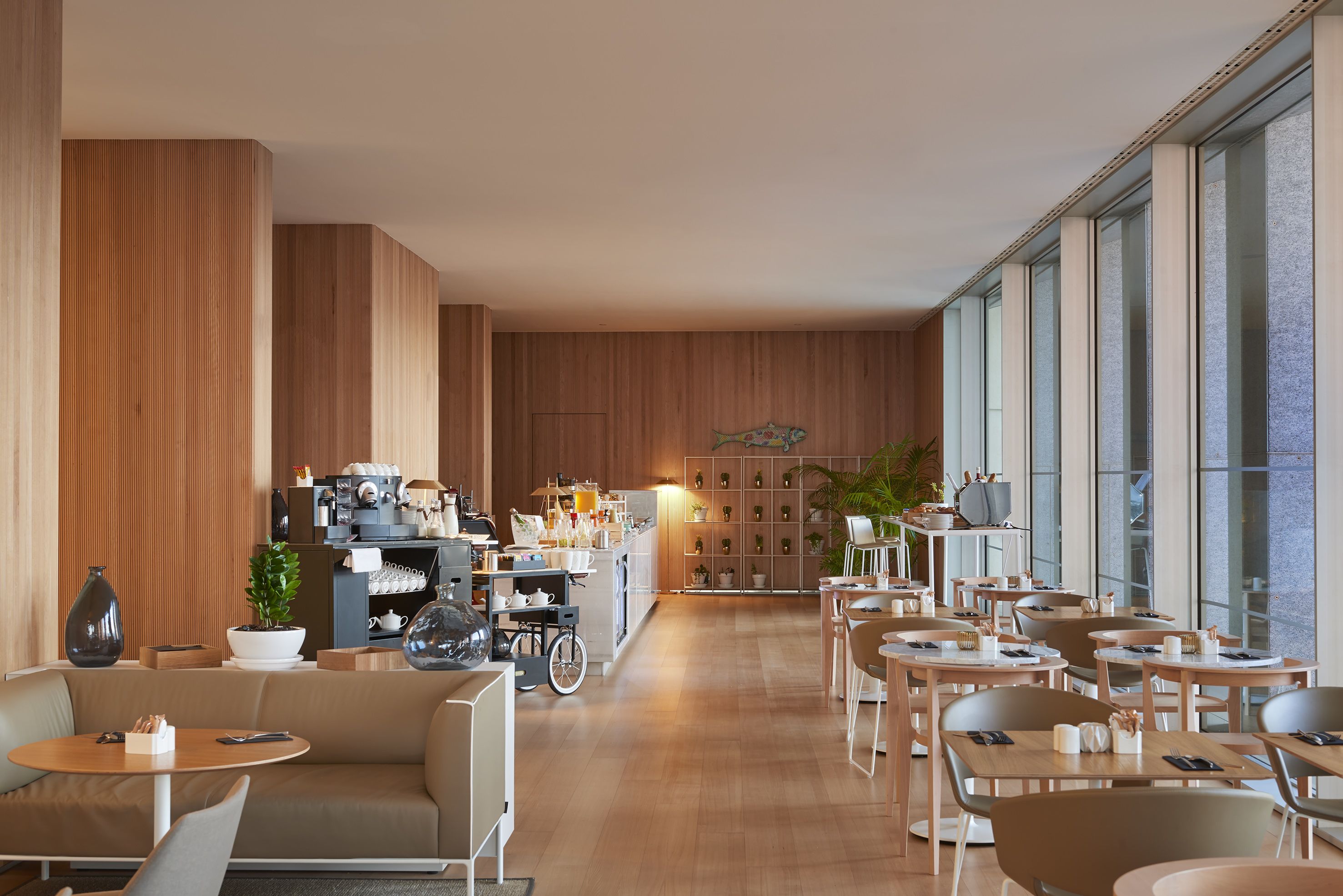 A modern cafe with wooden walls, a central counter with coffee machines, and multiple seating areas with tables and chairs.