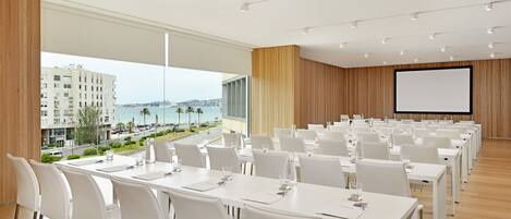 A conference room with white tables and chairs, a large screen, and a view of the sea and buildings.