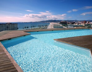 A swimming pool with wooden decks, a view of the sea, and distant buildings. - Melia Palma Bay (Palma de Mallorca)