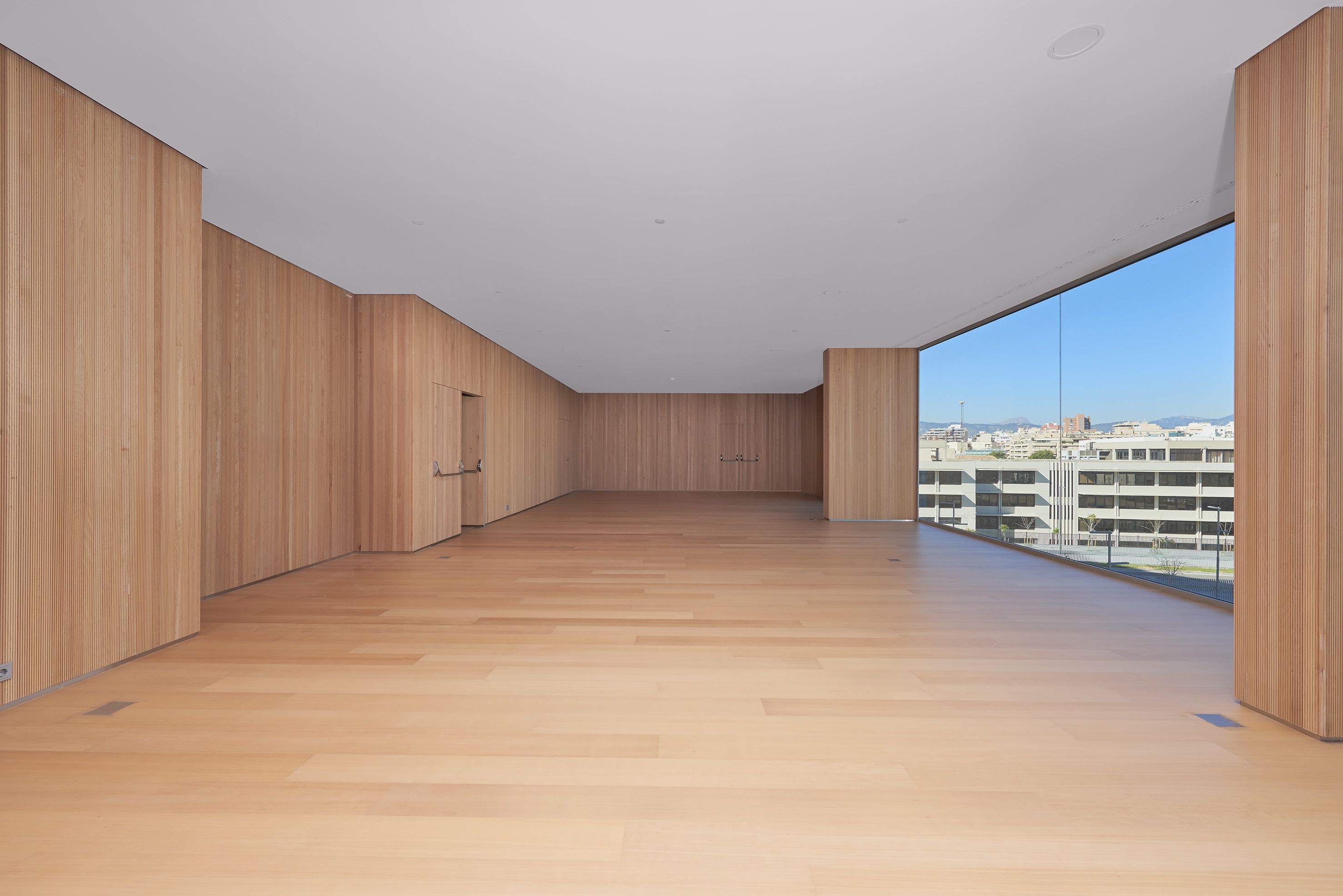 Amplia habitación con paredes de madera y un gran ventanal que ofrece vistas de edificios y un cielo despejado.