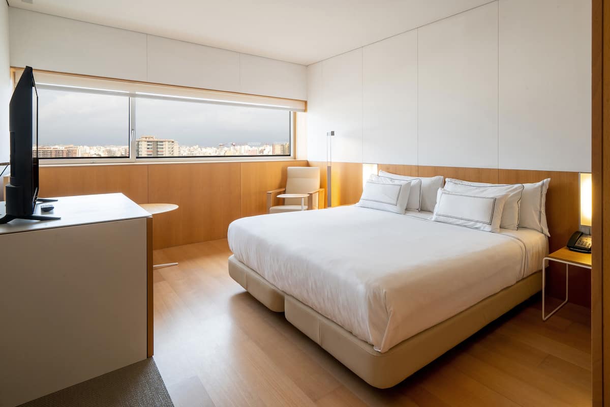 Quarto de hotel moderno com uma cama grande, uma escrivaninha, uma cadeira e vista para a cidade.