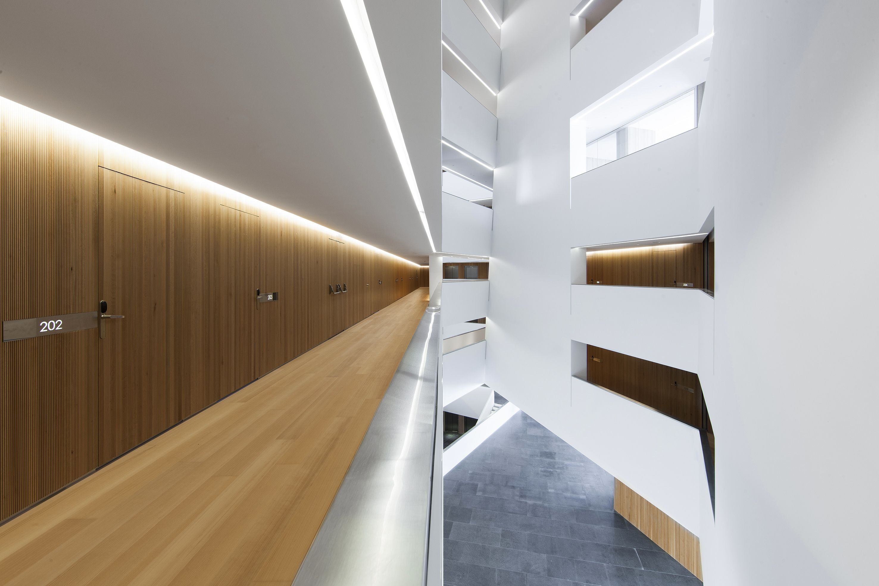 A modern hotel corridor with wooden paneling, numbered doors, and a central staircase.