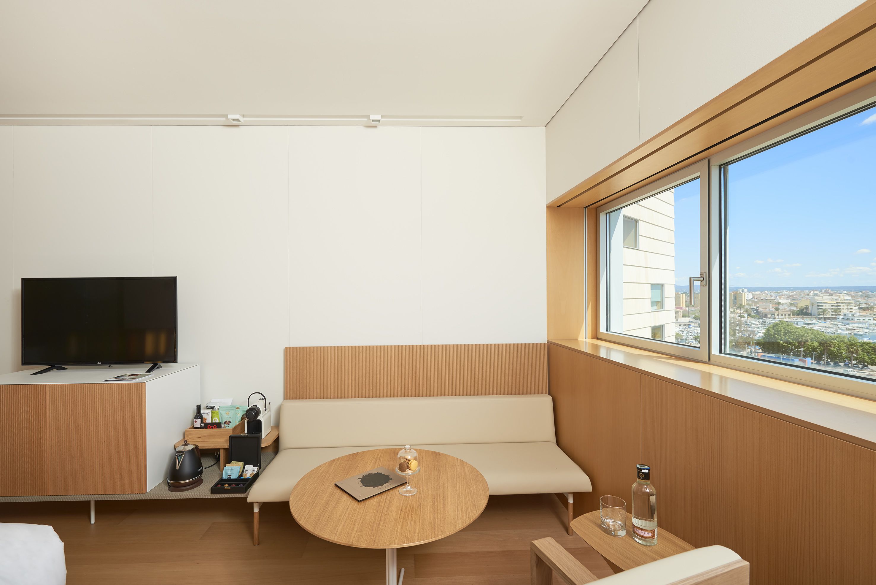 Una habitación de hotel moderna con un ventanal grande, un televisor de pantalla plana, una mesa de madera, un sofá y una pequeña zona de kitchenette.