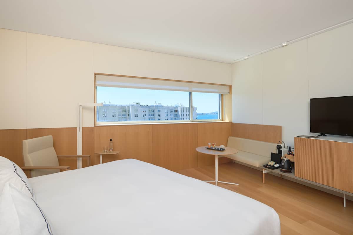 Quarto de hotel com uma cama grande, um sofá, uma mesinha, uma televisão e uma janela com vista para a cidade.