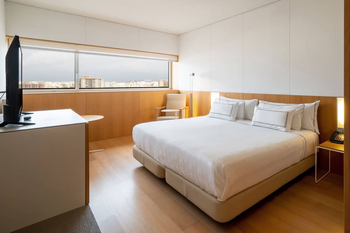Quarto de hotel moderno com uma cama grande, uma escrivaninha com computador, uma cadeira e vista para a cidade.
