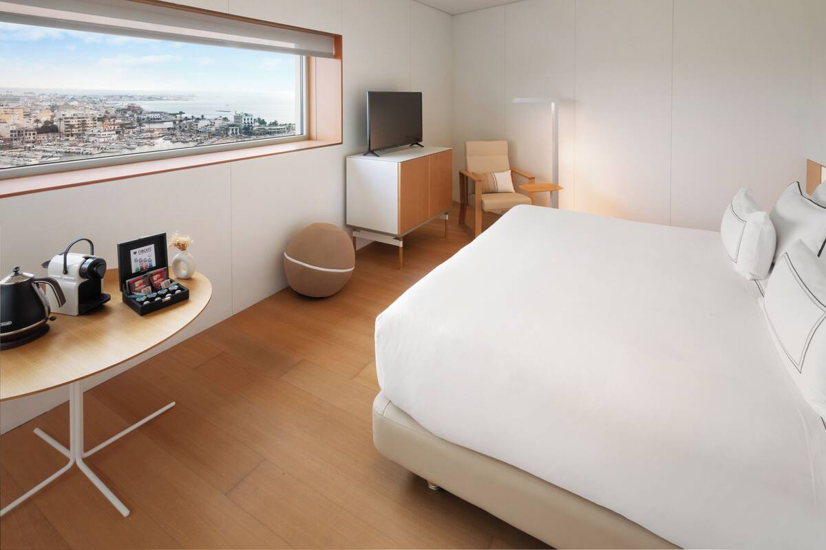 Quarto de hotel moderno com uma cama grande, uma escrivaninha com máquina de café, uma televisão sobre um móvel, uma cadeira e vista para a cidade através de uma janela ampla.