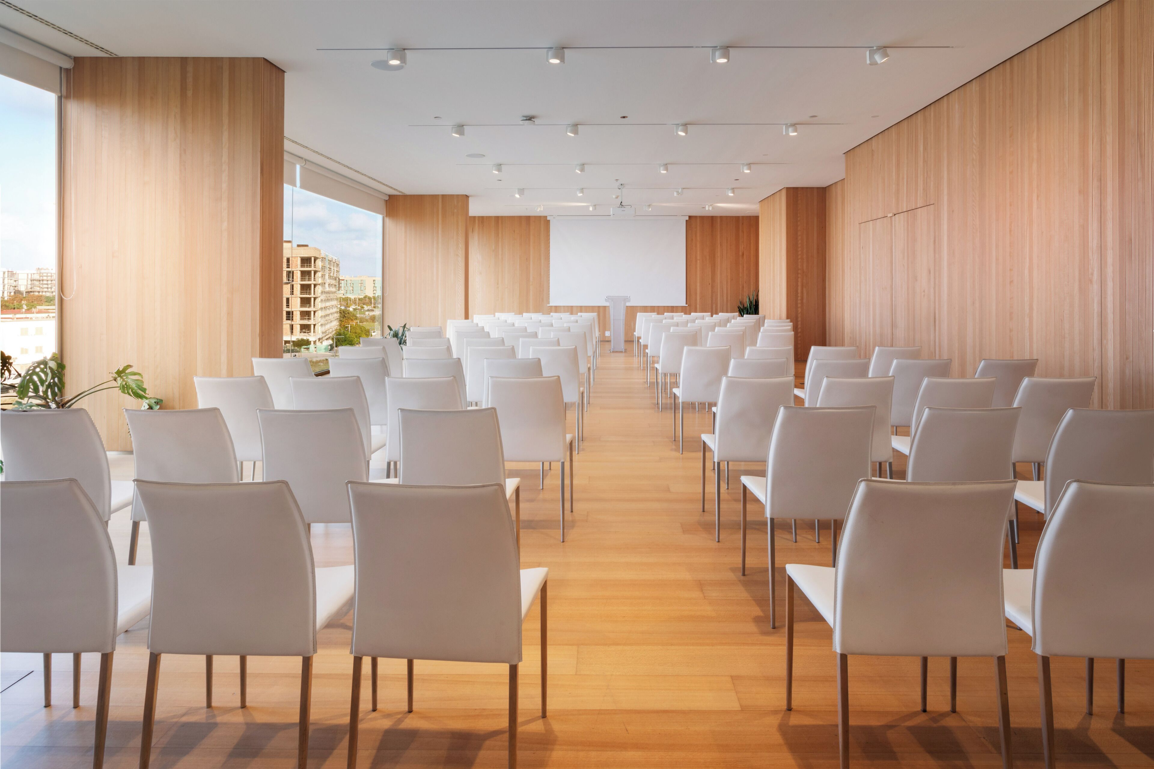 A modern conference room with rows of white chairs, wooden floors, and large windows offering a city view.